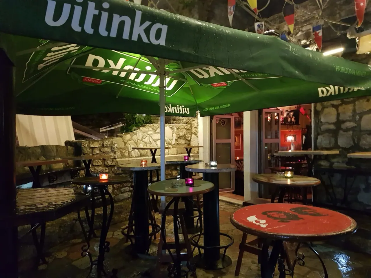 Candle-lit high tables and stools on the terrace of Jeff Bar in Budva's Old Town at night, under a green umbrella and strings of pennant flags against a stone wall