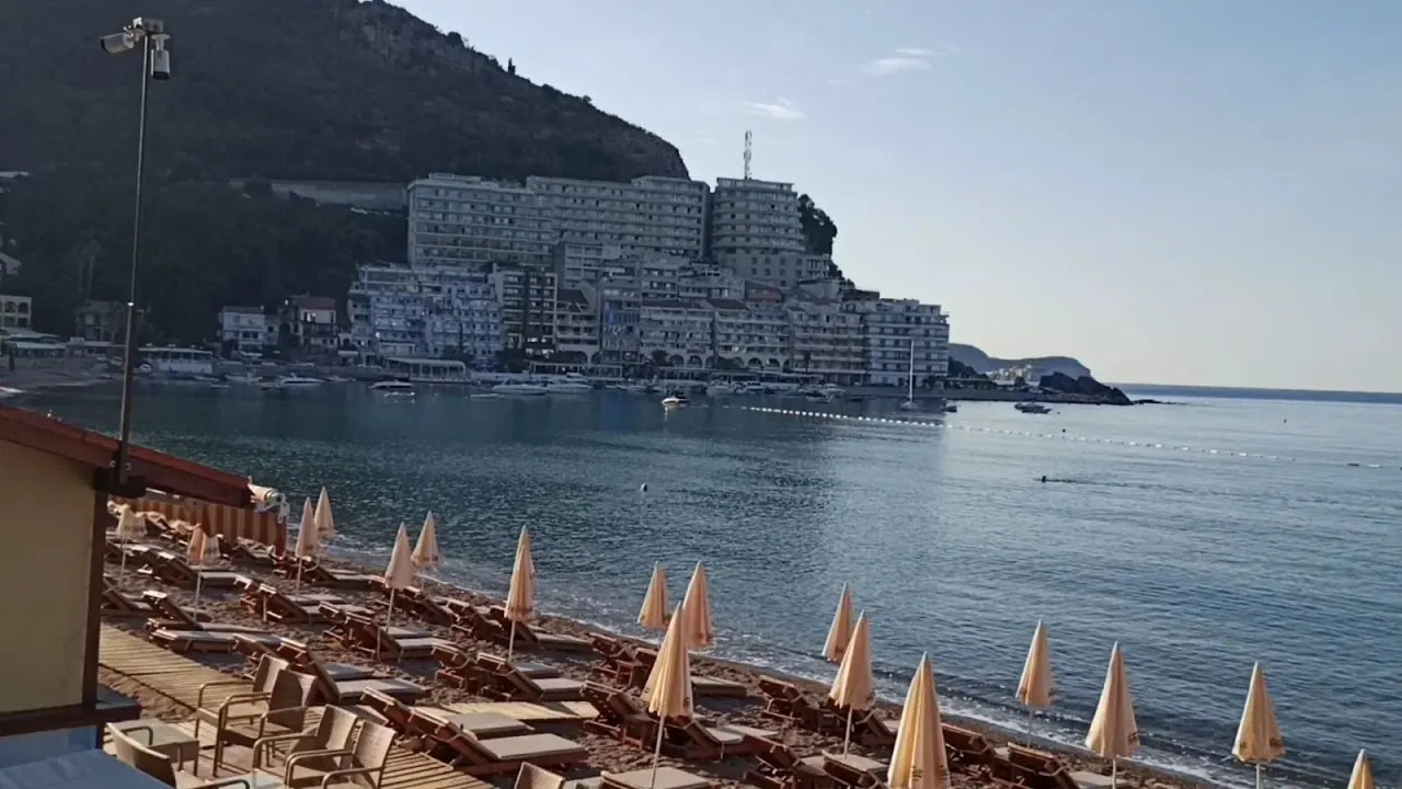 Empty loungers and folded parasols on Bečići Beach in the early morning, looking across the calm bay to the tiered hillside apartments of Rafailovići