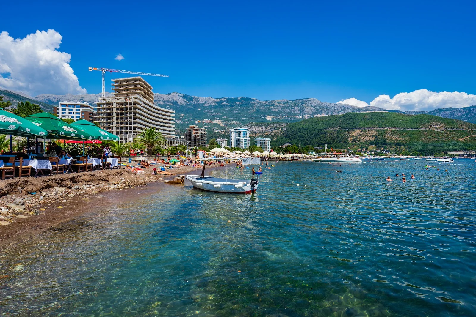 Shaded tables and green umbrellas of Beach Greco on the sand at Budva, with a wooden boat moored offshore and mountains behind the bay