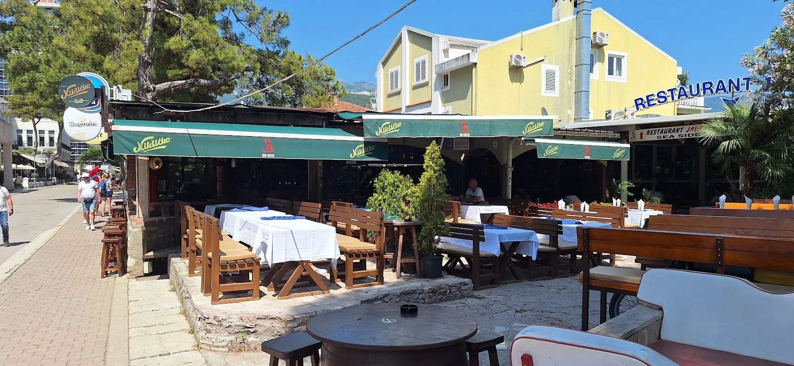Green Nikšićko-branded awnings over the street-side terrace of Beer & Bike Club in Budva, with wooden benches and bar stools along the promenade