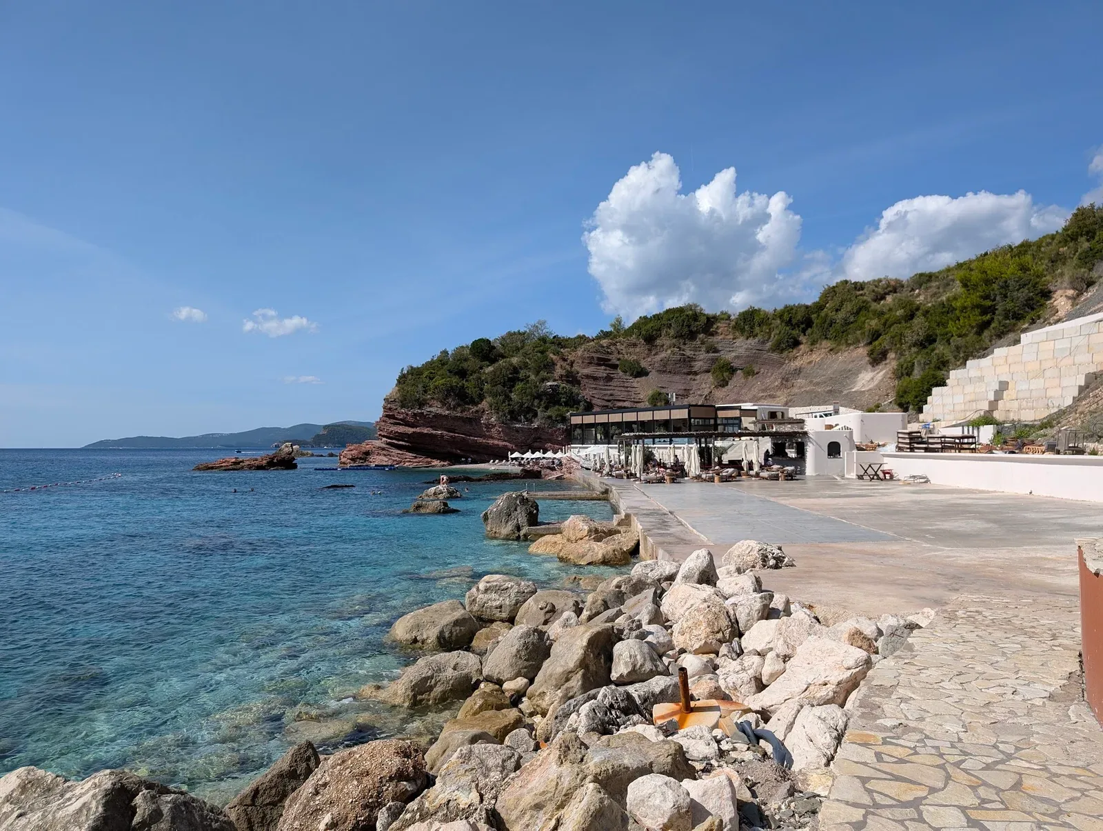 The white beach club pavilion at Galija Beach below a red rock headland, with turquoise water and a stone sun terrace along the shore