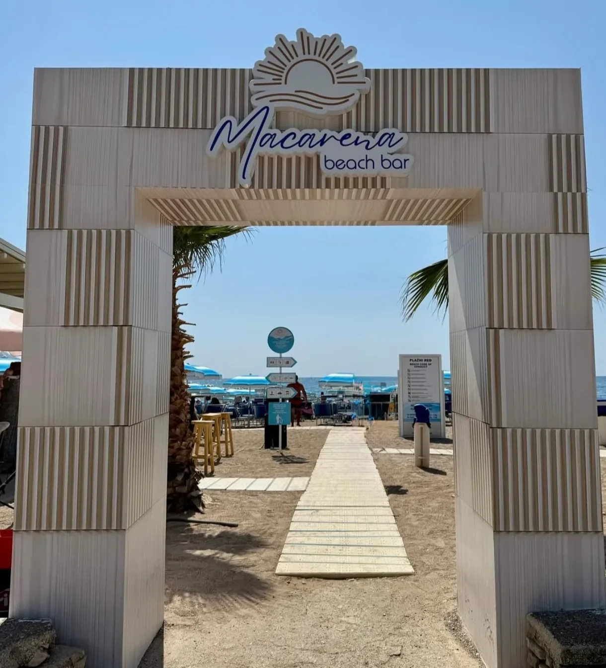 The pale stone entrance arch of Macarena beach bar in Bečići, with a boardwalk running through palms to blue umbrellas on the sand