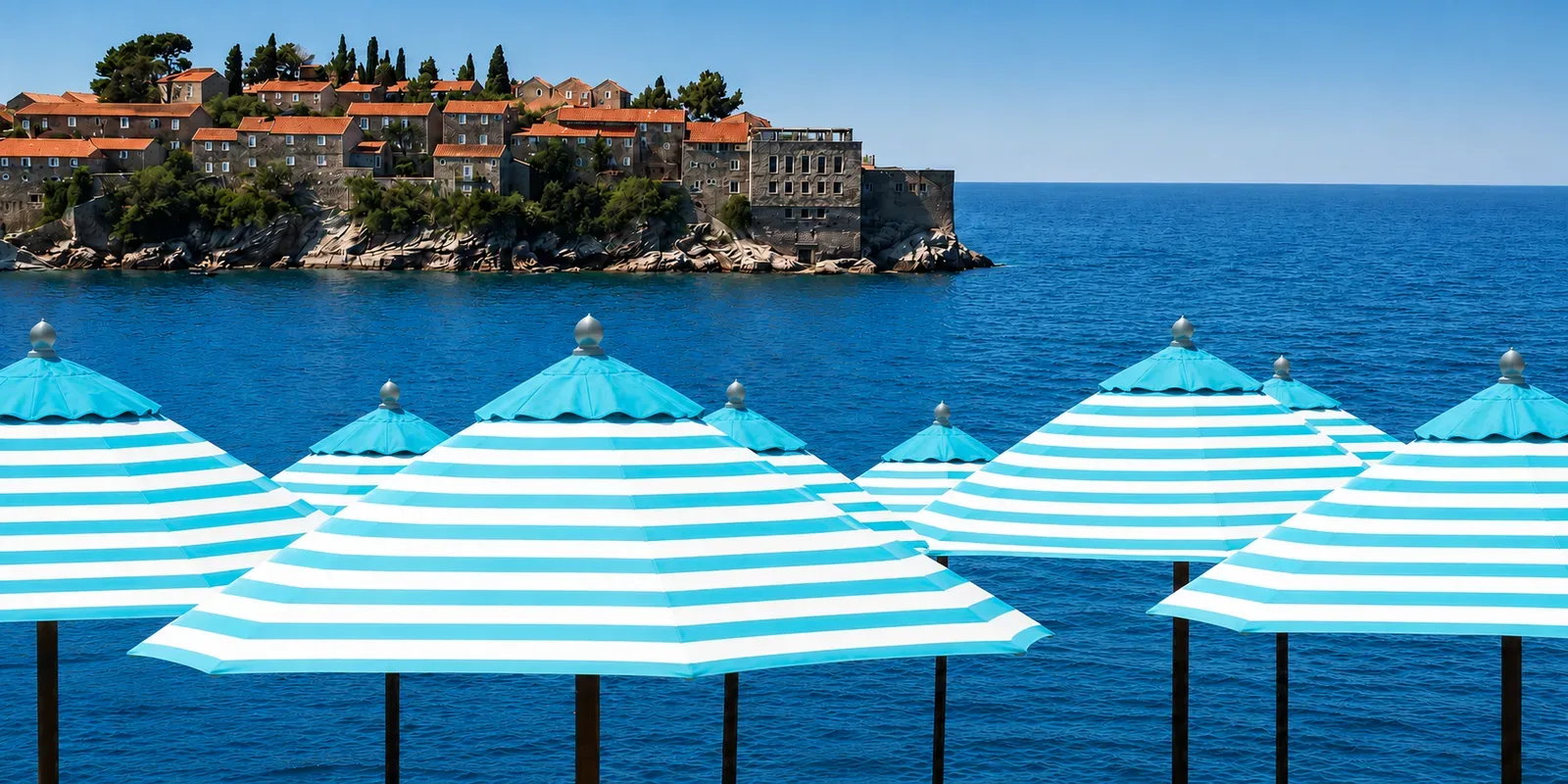 Rows of turquoise and white striped umbrellas at Nammos Montenegro, with the walled islet of Sveti Stefan across the blue water behind
