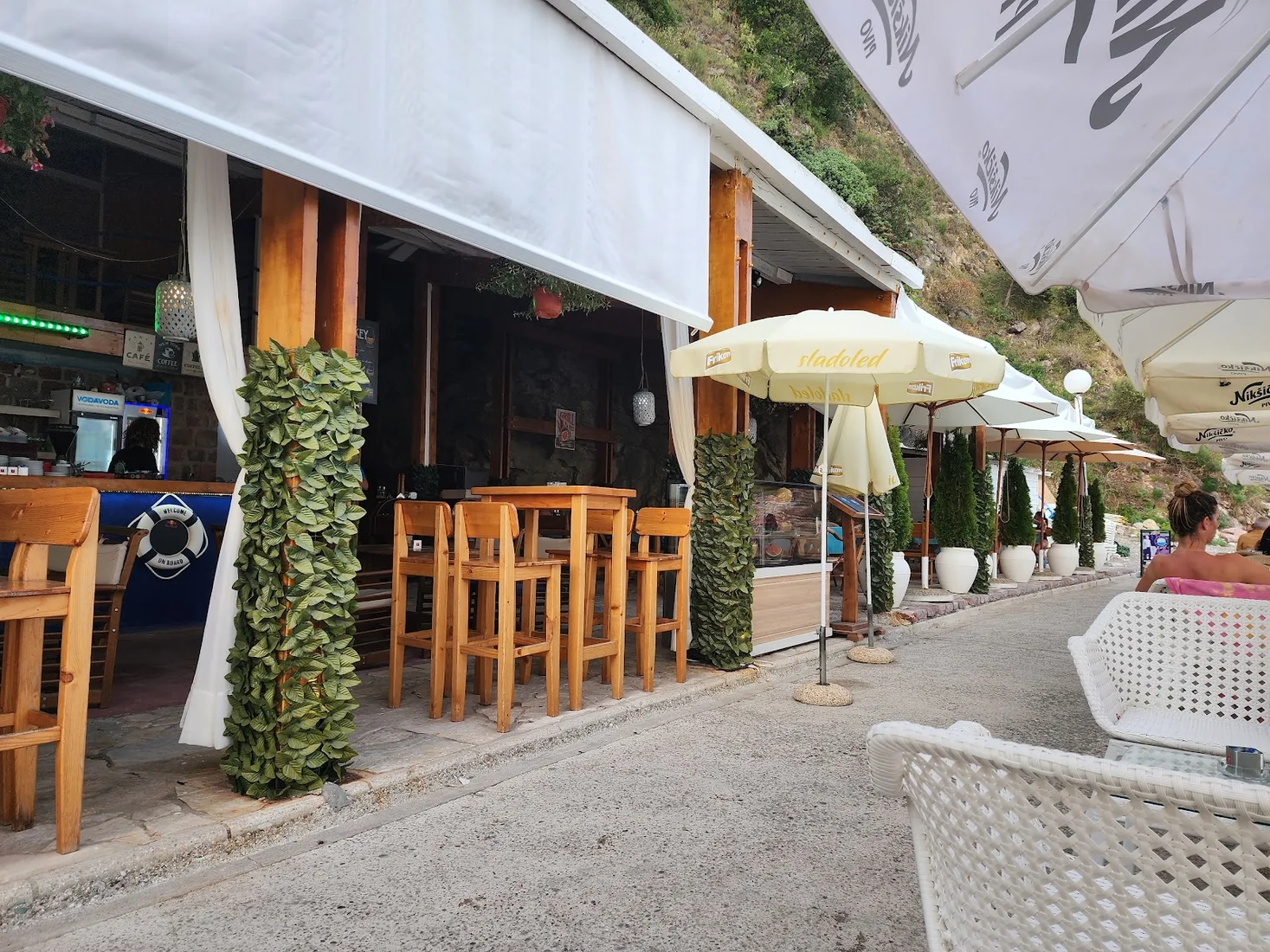 Wooden bar counter, stools and white awnings on the seafront path at Stijene Lounge Bar in Rafailovići, with umbrellas and a rocky hillside behind