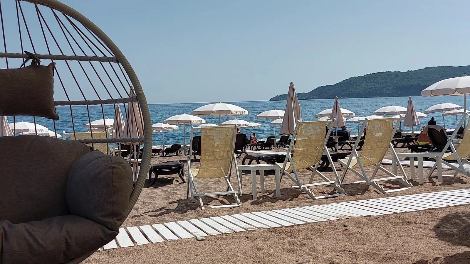 A hanging egg chair, sun loungers and white umbrellas on the sand at Sunset Beach Bar in Bečići, looking out to the Adriatic and a green headland