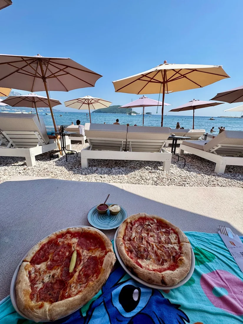 Beige umbrellas and padded loungers on the pebble beach at Torch Beach Club in Budva, looking out to Sveti Nikola island, with two pizzas on a towel in the foreground
