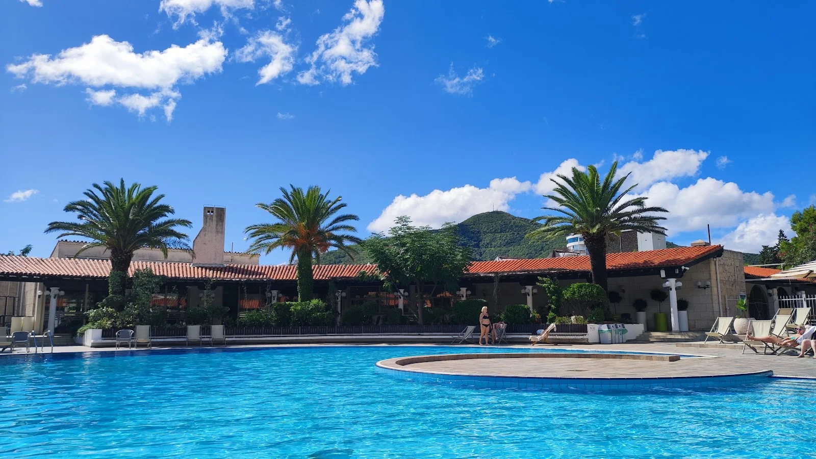 Large outdoor pool ringed by palm trees and terracotta-roofed buildings at Hotel Slovenska Plaža in Budva