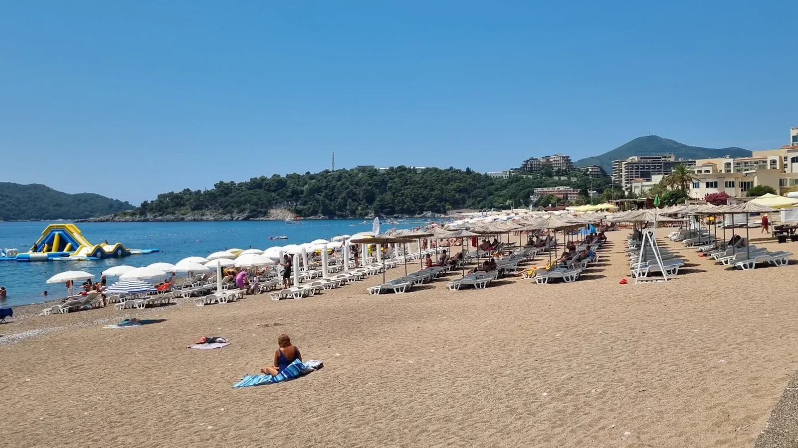Rows of sunbeds and thatched umbrellas on the sandy strand of Bečići Beach, with an inflatable water park in the bay and a wooded headland beyond