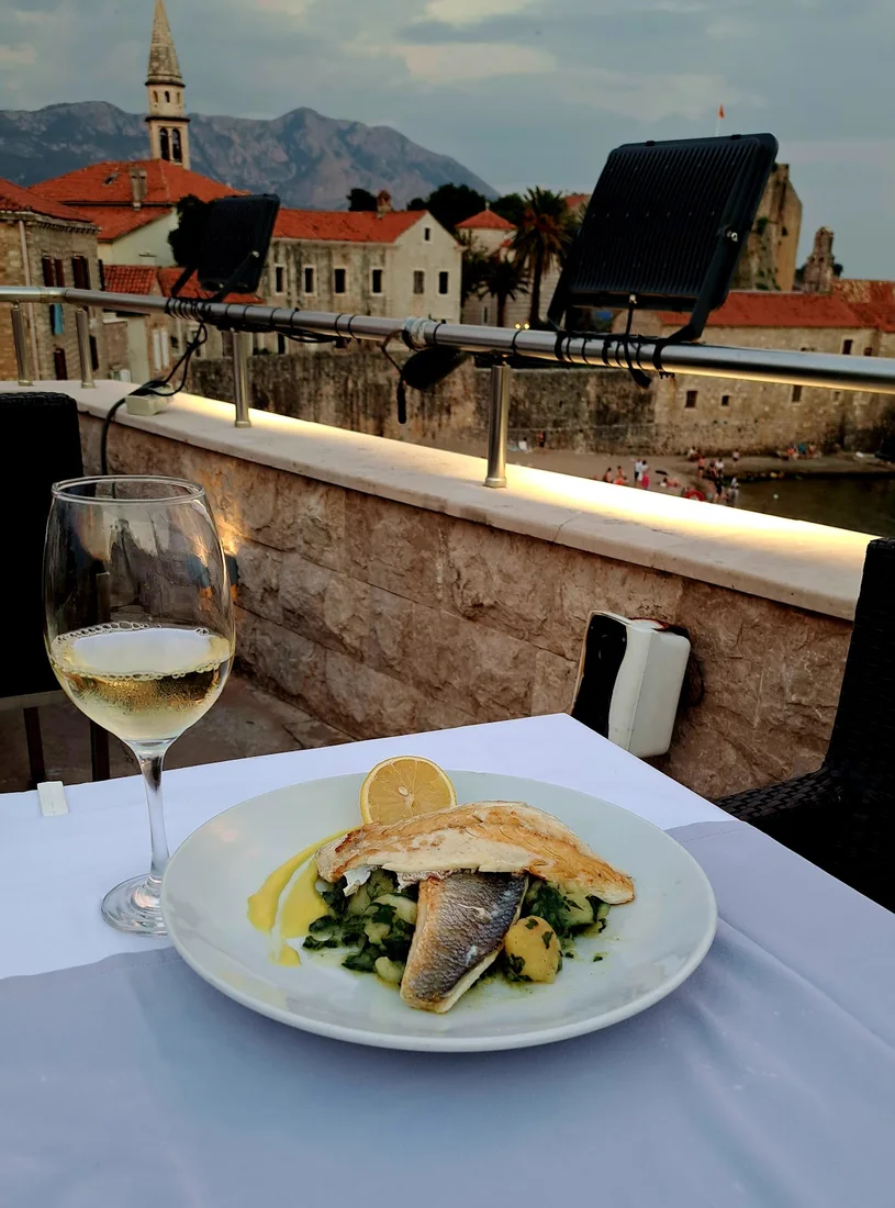 Plated grilled fish and a glass of white wine on a rooftop table overlooking Budva's Old Town roofs and church tower at dusk