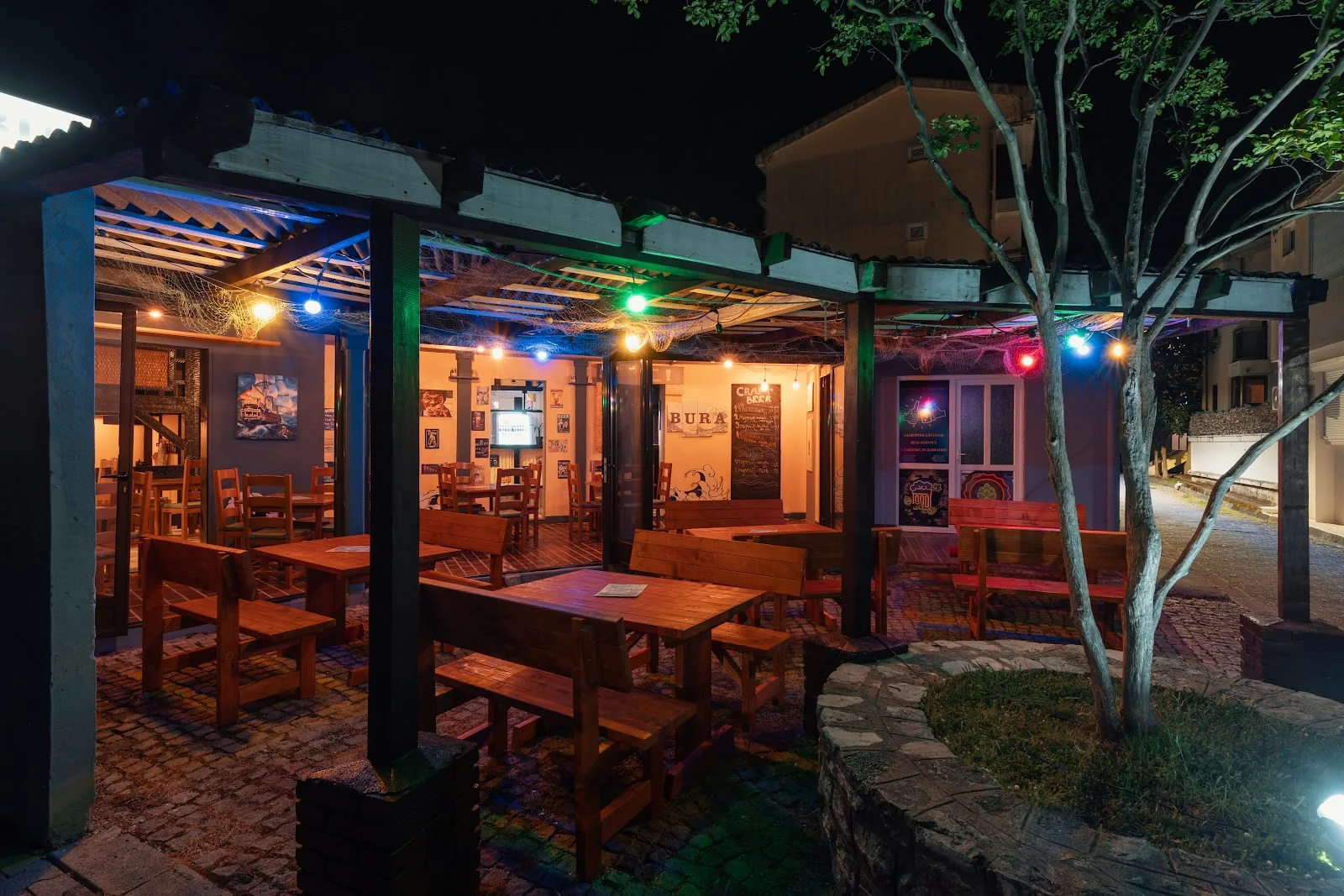 String-lit courtyard with wooden benches at night under the BURA sign at Bura Pub in Budva