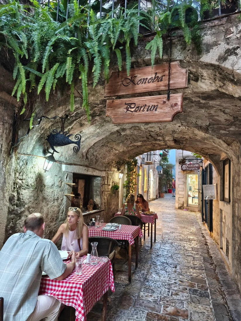 Red-checked tables under the vaulted stone arch and hanging ferns of Konoba Portun in Budva's Old Town