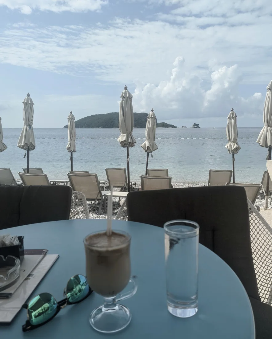 A turquoise-topped table with an iced coffee looking out over beach umbrellas and Sveti Nikola island at La Bocca in Budva