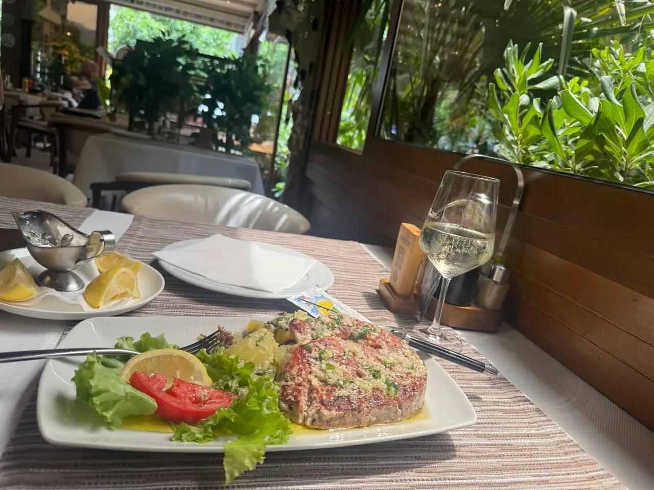 A grilled pork chop with salad and a glass of white wine on a garden-side table at Restaurant Adriatic in Budva